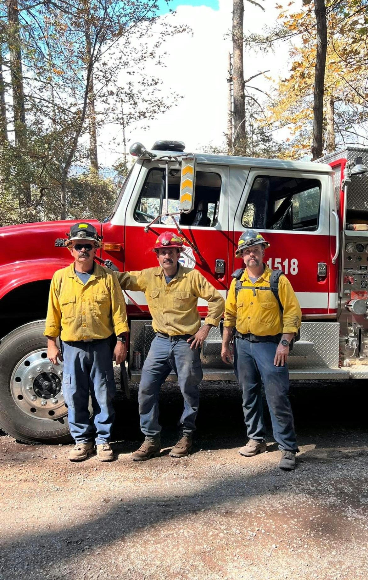 Three firefighters in yellow shirts and helmets in front of a red fire truck, surrounded by trees under a blue sky.