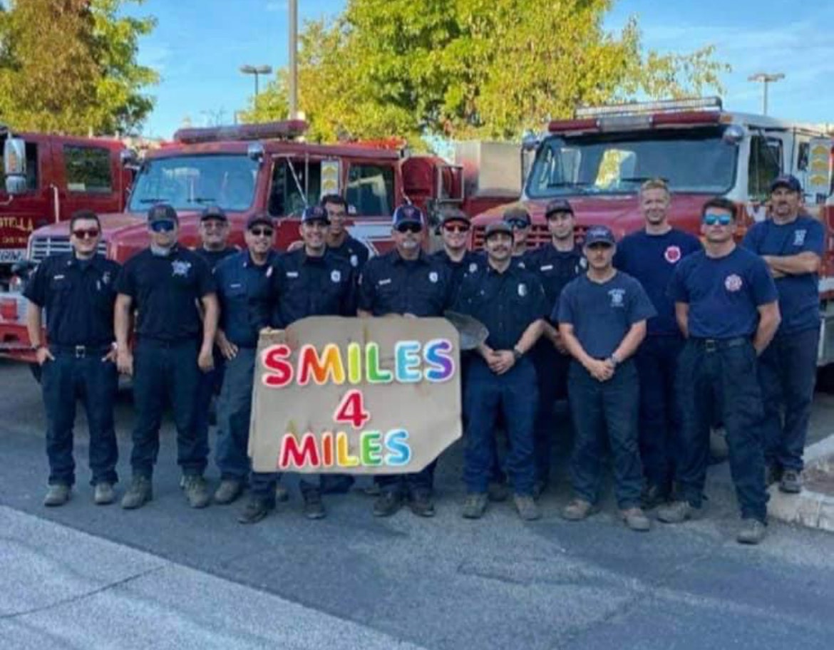 Group of firefighters posing with a "SMILES 4 MILES" banner in front of fire trucks.