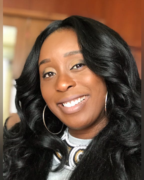 A smiling person with long, curly hair and hoop earrings, wearing a decorated top, in a warm indoor setting.