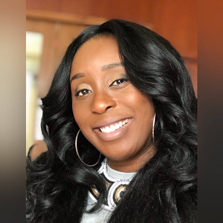 A smiling person with long, curly hair and hoop earrings, wearing a decorated top, in a warm indoor setting.