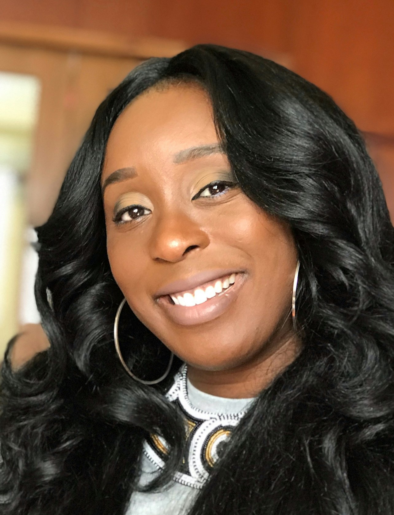 A smiling person with long, curly hair and hoop earrings, wearing a decorated top, in a warm indoor setting.
