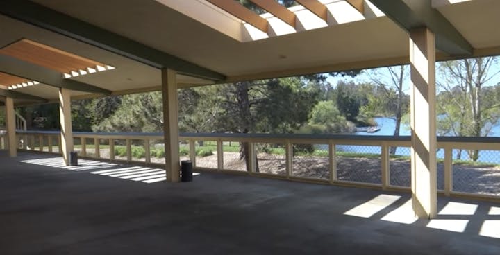 The image shows a spacious, well-lit area with a view of a lake and trees, featuring beams and a railing.