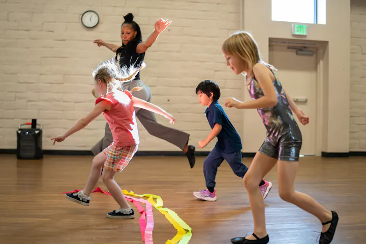 A group of children are dancing in a studio, guided by an instructor. Colorful streamers are scattered on the floor.