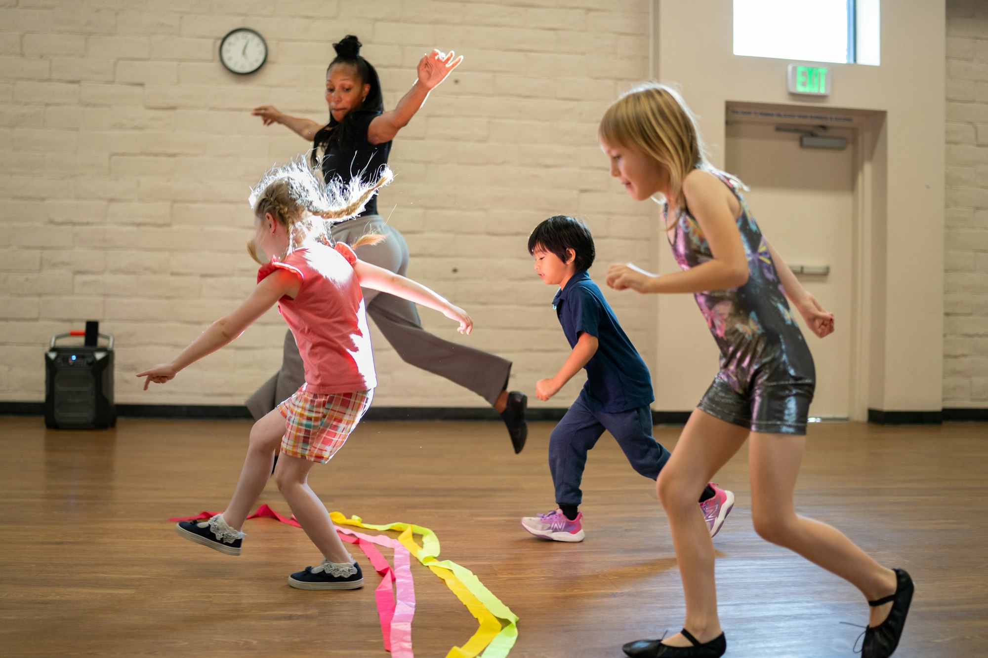 A group of children are dancing in a studio, guided by an instructor. Colorful streamers are scattered on the floor.