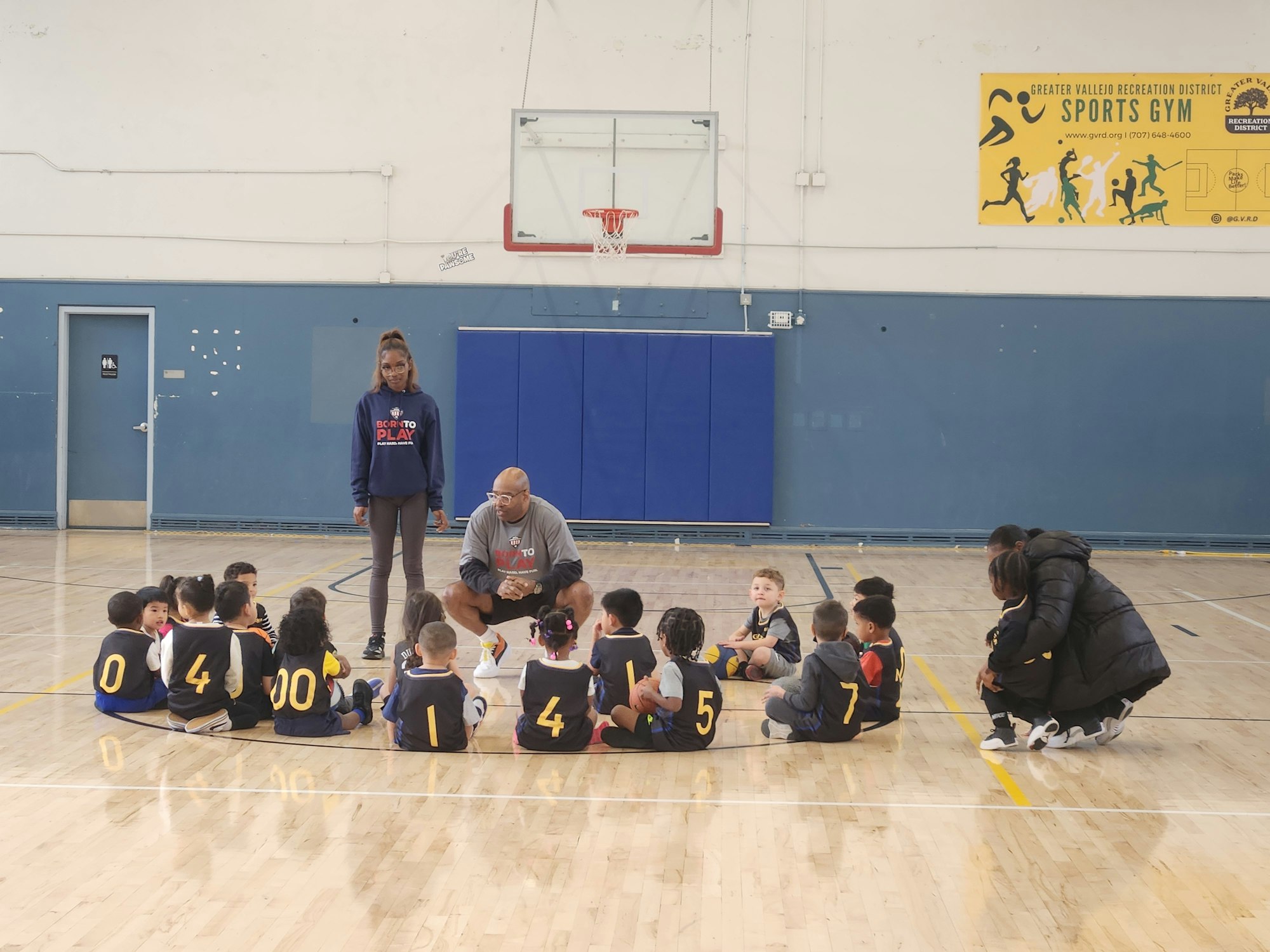 A group of young children in sports jerseys sits on a gym floor, listening to a coach and an instructor during practice.