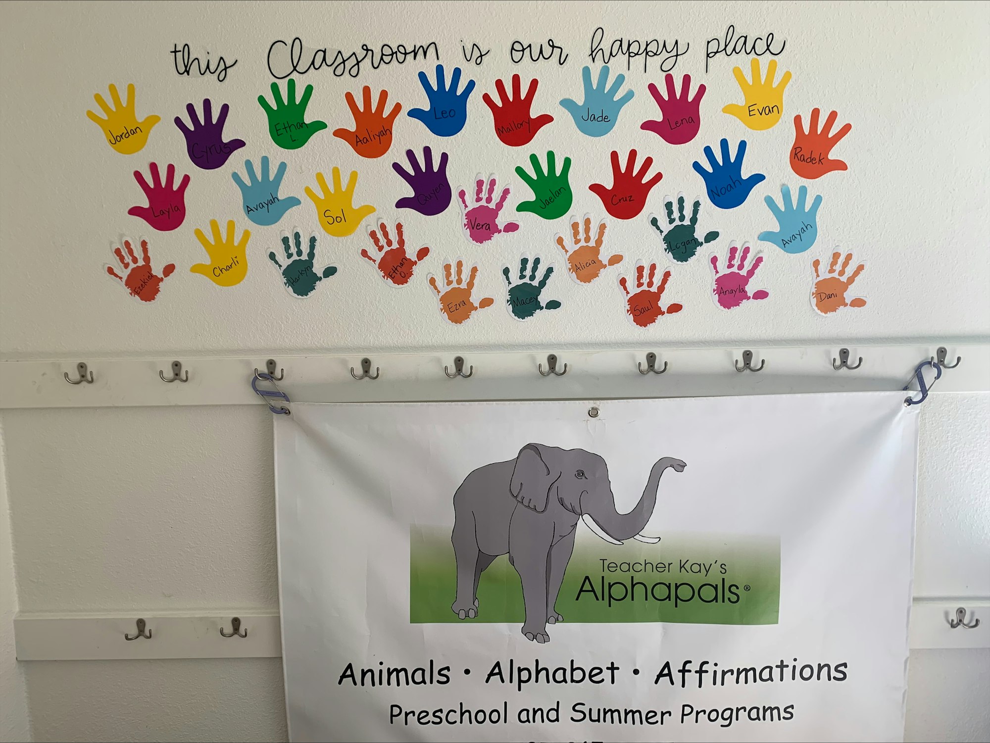 Colorful handprints with names on a wall above a sign mentioning animals, alphabet, affirmations for preschool and summer programs.