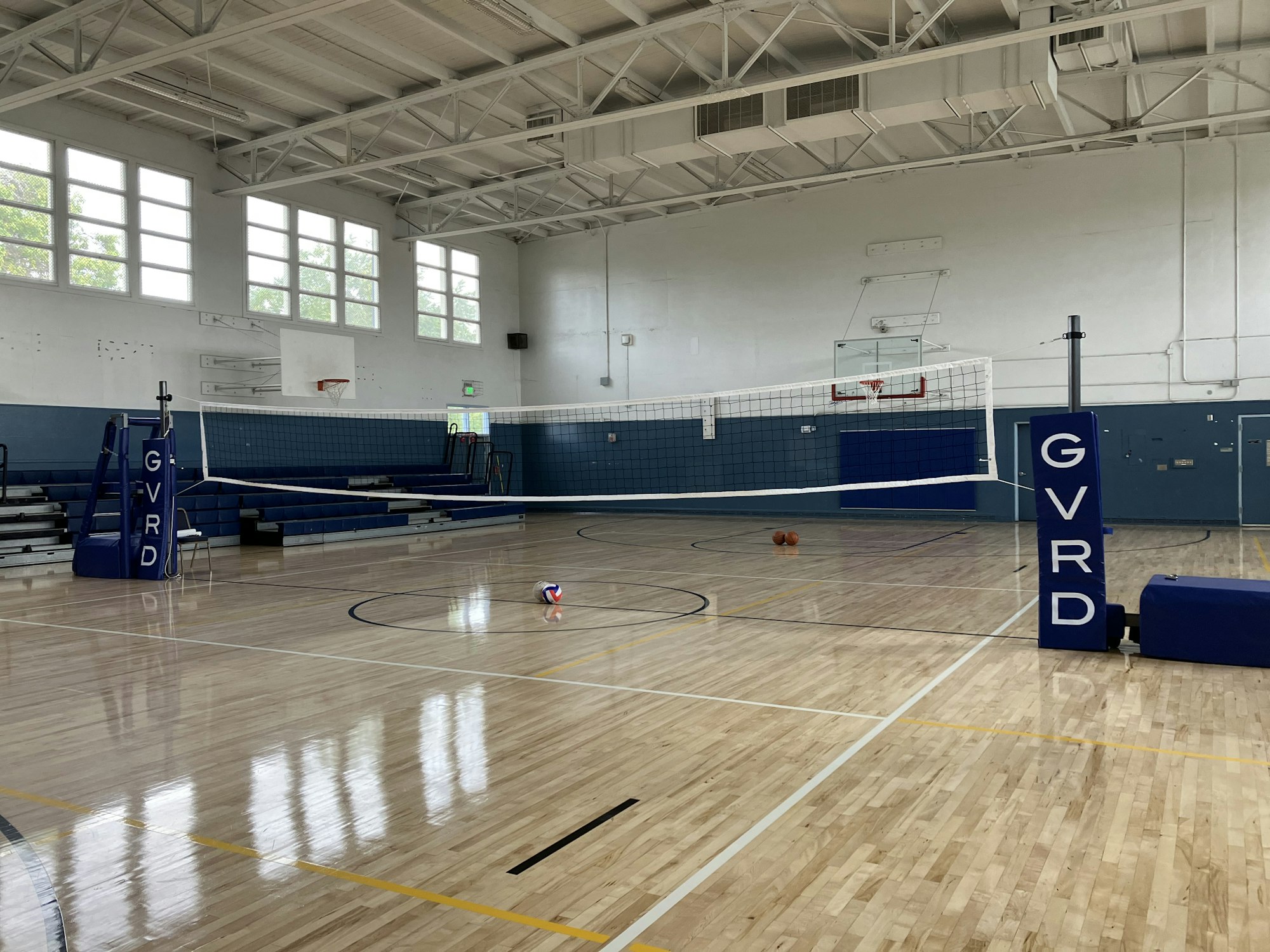 Indoor gym with volleyball net, bleachers, basketball hoop, and scattered balls on the floor.