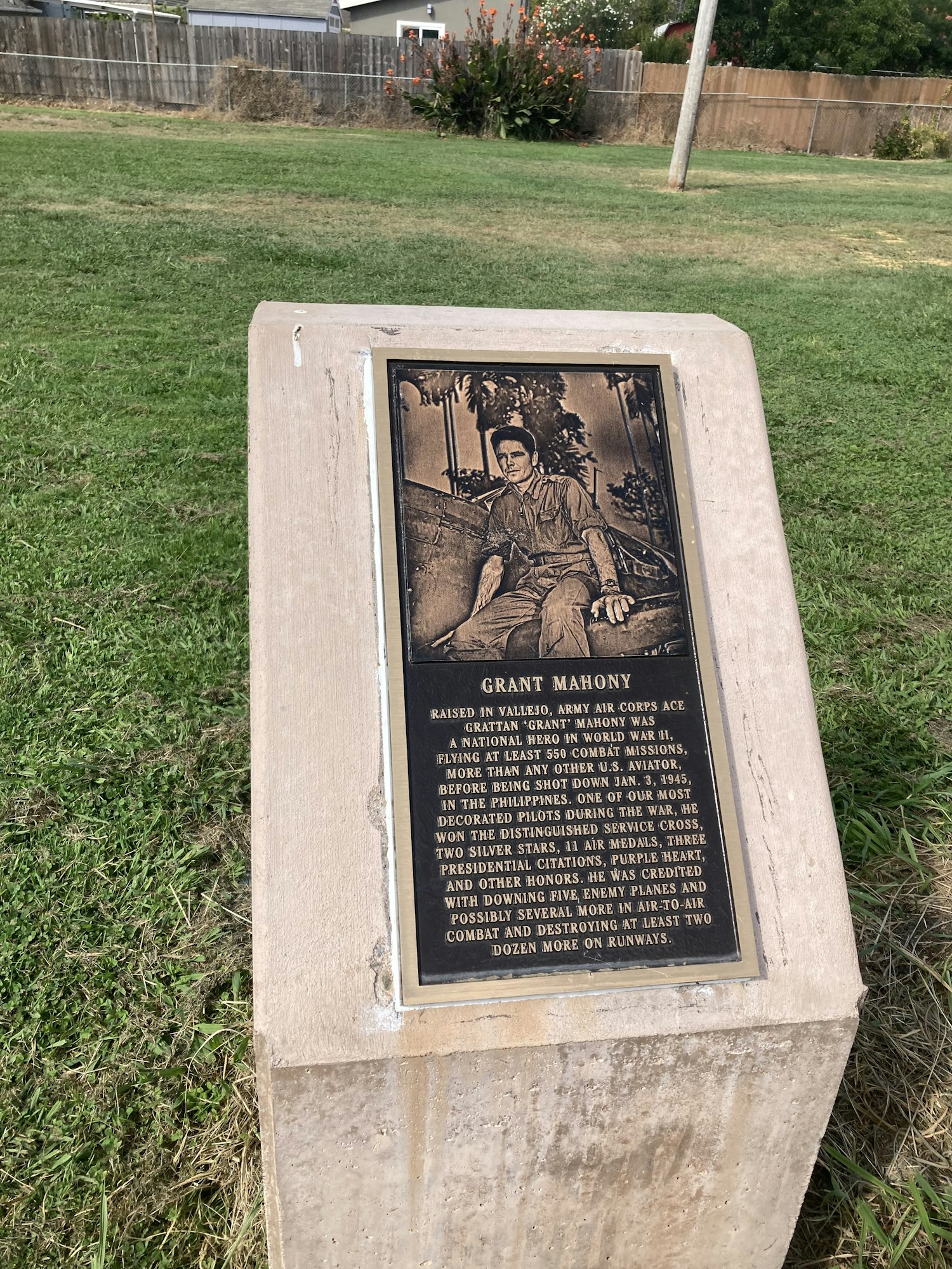 An angled photo of a commemorative plaque with engraved text and image, mounted on a concrete stand in grassy surroundings.