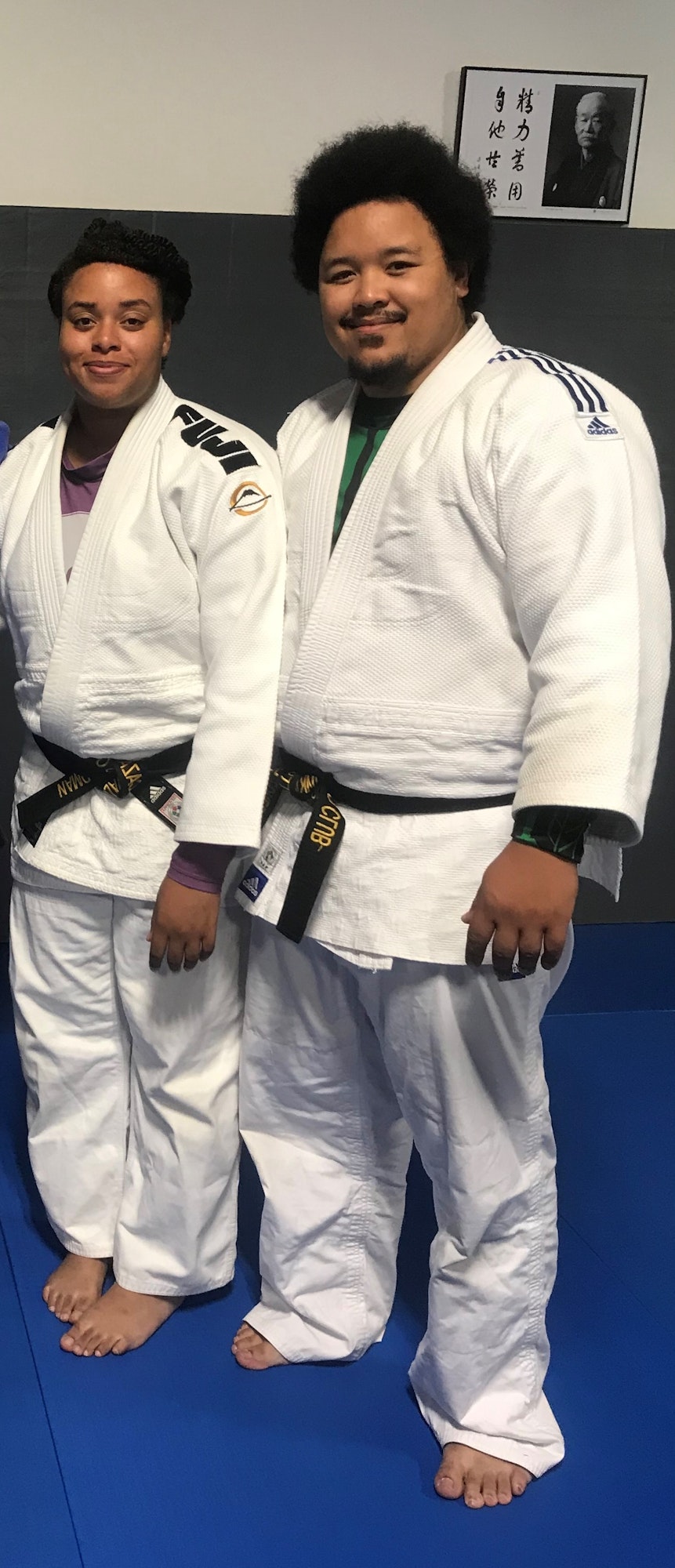 Two individuals in judo gis with black belts, standing on a blue mat, with a picture on the wall behind them.