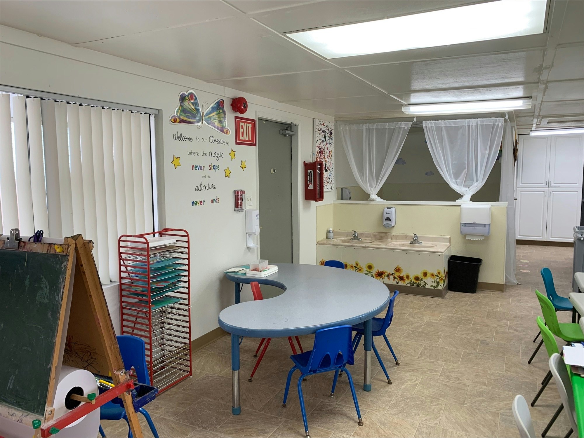 A classroom with a chalkboard, child-sized tables and chairs, drawings, educational decor, and a handwashing station.