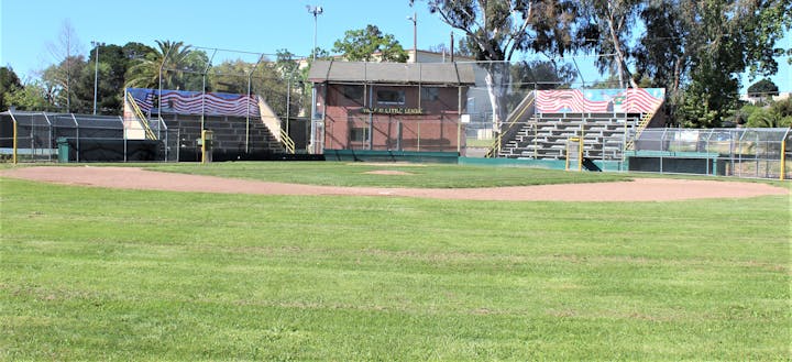 A baseball field with a grassy infield, a backstop, and small bleachers in the background, featuring a brick building nearby.