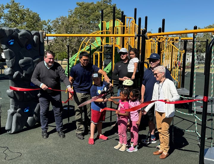 A group of people are cutting a red ribbon at a playground's opening ceremony.