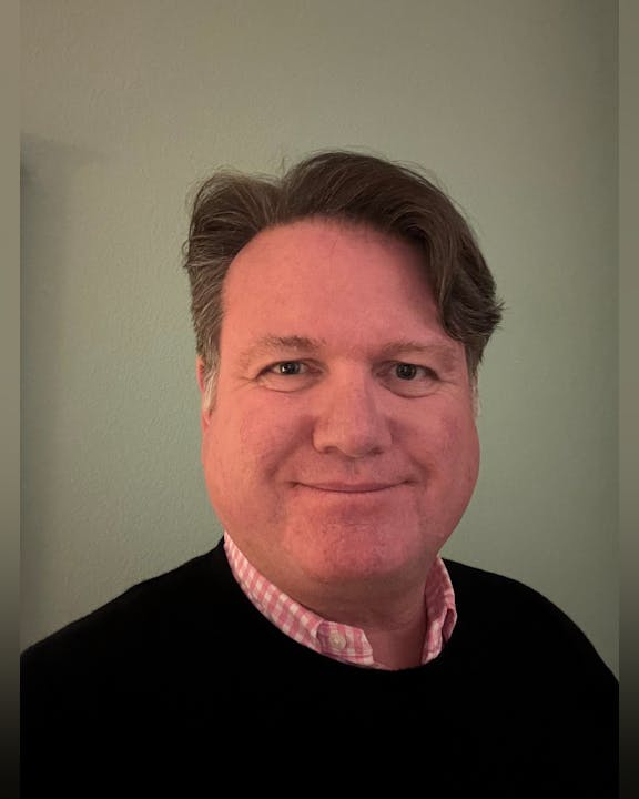 General Manager Gabe Lanusse. A person with short brown hair and a pink checked shirt under a black sweater, smiling in front of a plain background.