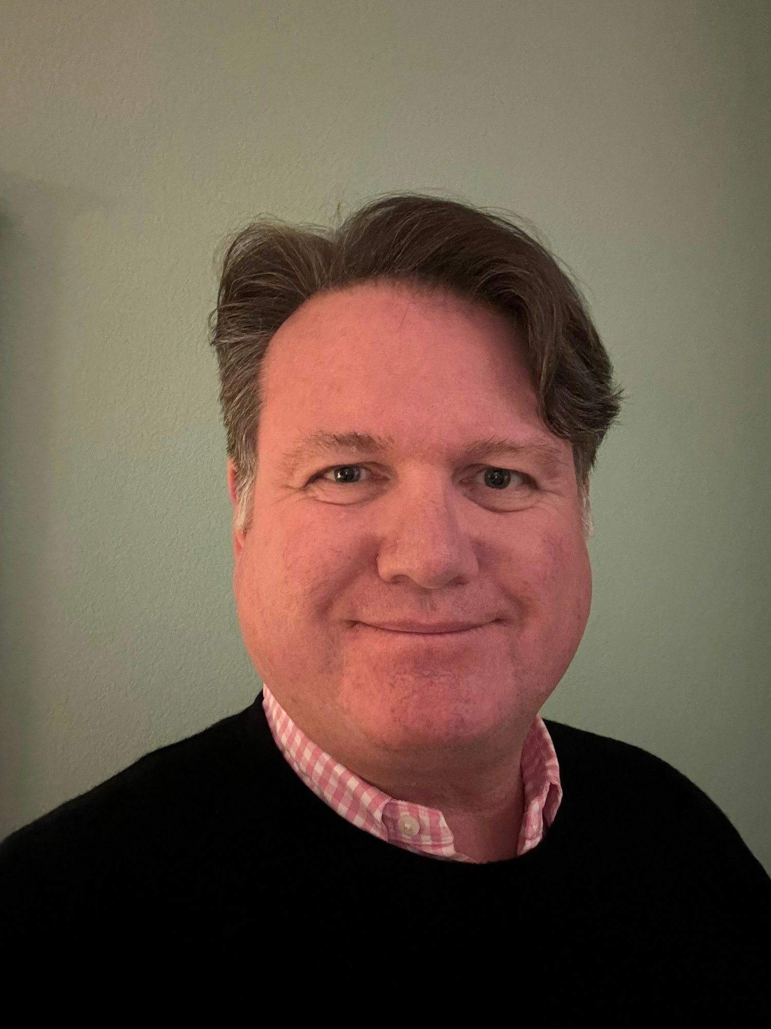 General Manager Gabe Lanusse. A person with short brown hair and a pink checked shirt under a black sweater, smiling in front of a plain background.