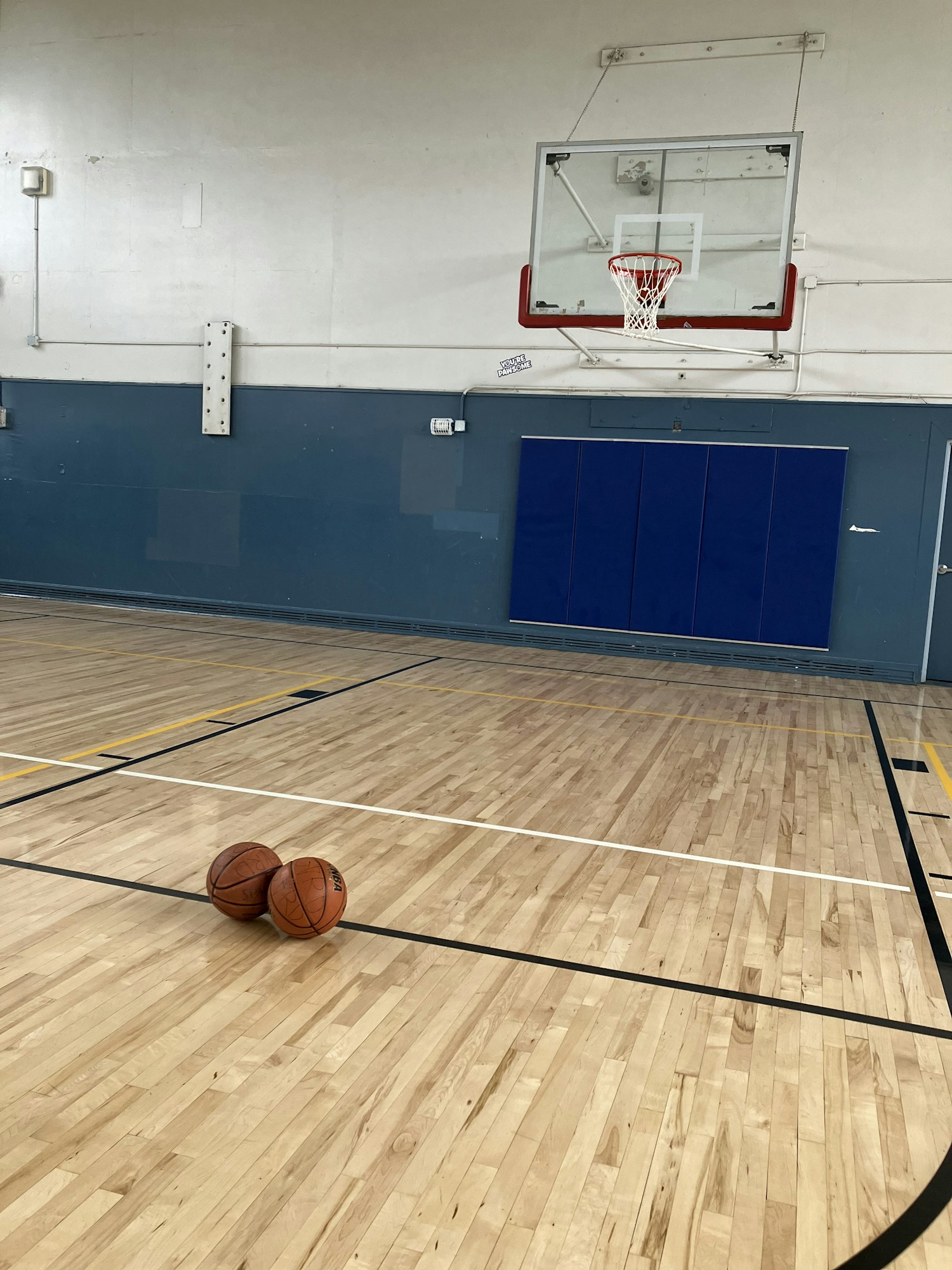 A basketball and hoop in a gym, with a reflection on the glossy wooden floor.