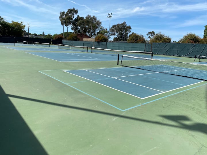 A set of empty tennis courts under clear skies, with blue and green surfaces and netting.
