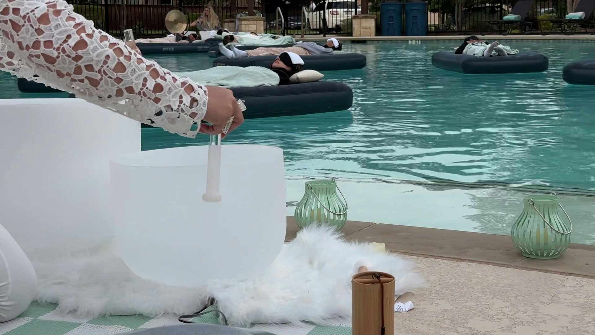 A serene poolside setting with people relaxing on floats, while someone prepares a crystal singing bowl for a wellness session.