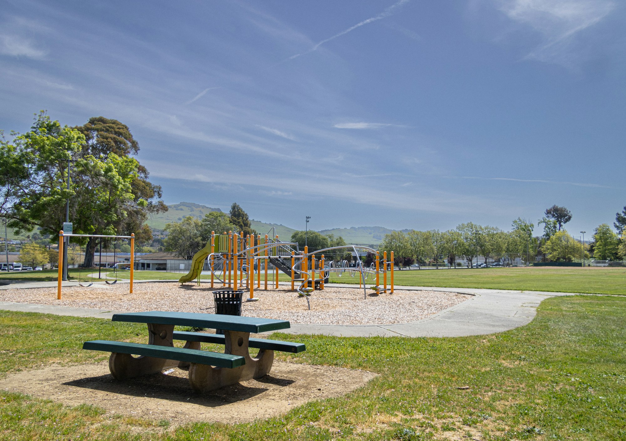A park with a picnic table, playground equipment, and distant hills under a blue sky.