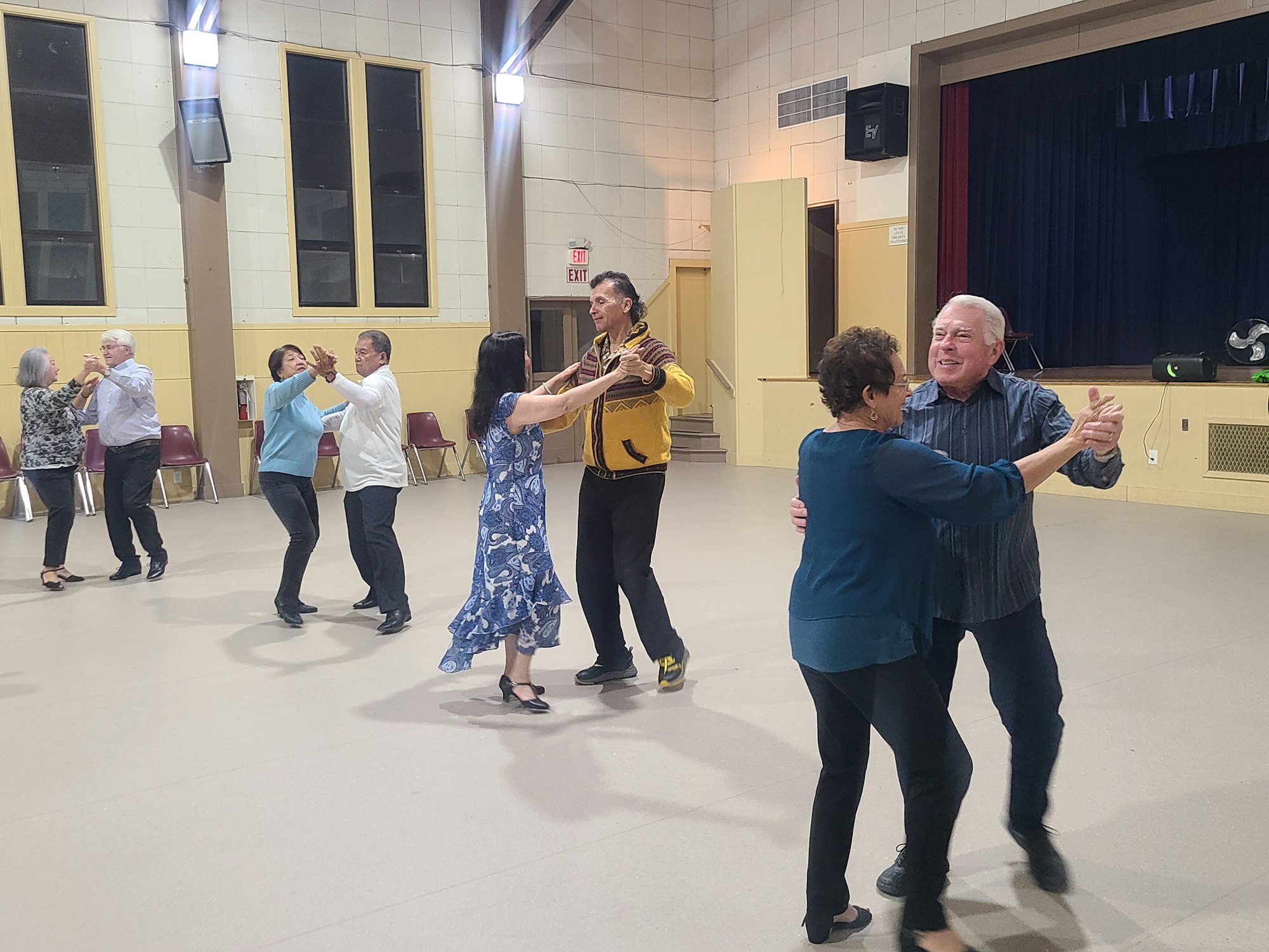 A group of couples are dancing in a large hall with chairs along the walls.
