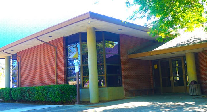 The image shows a modern brick building with large windows, a flat roof, and green landscaping in front.