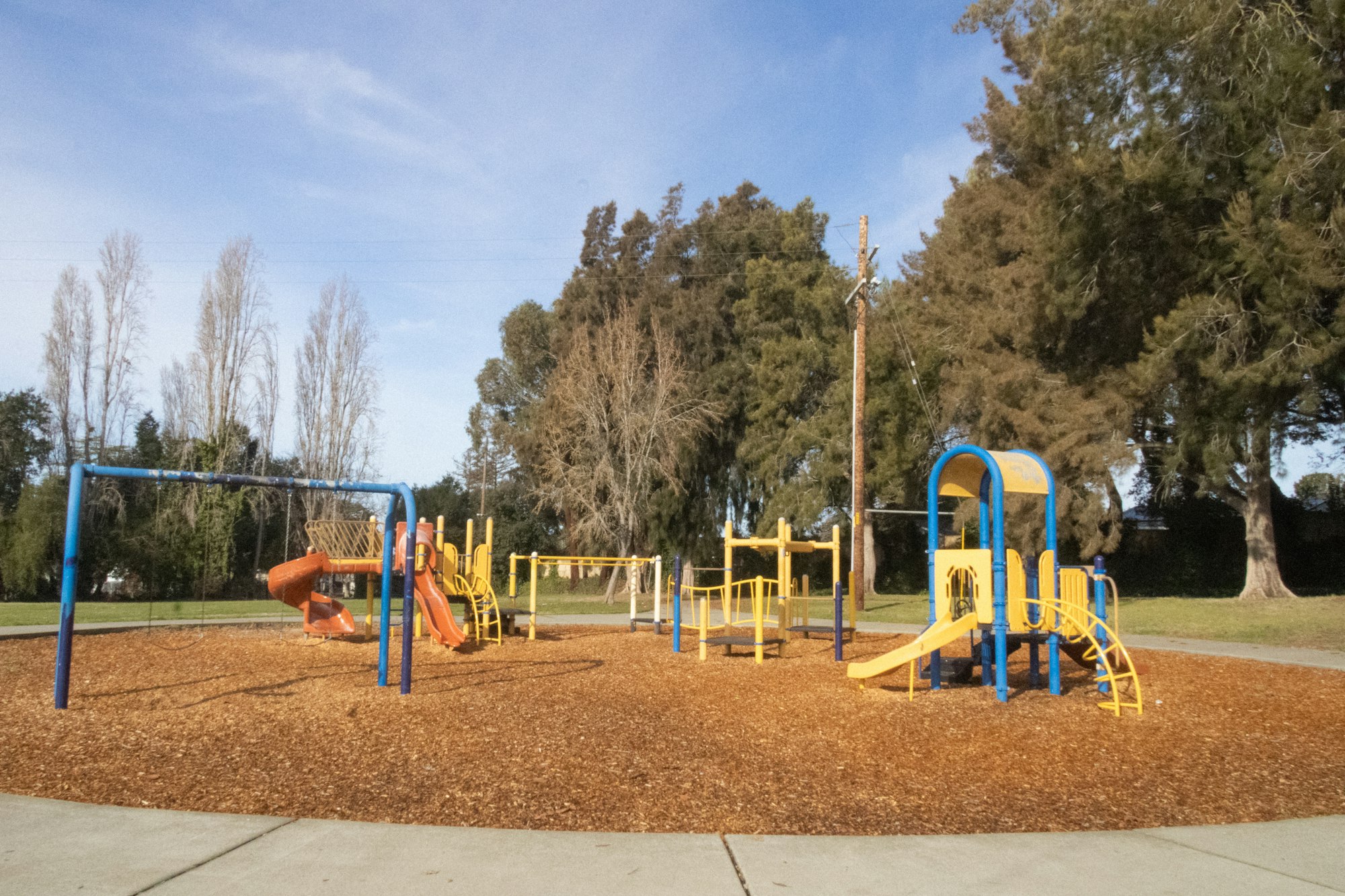A colorful playground with slides and swings on a sunny day, surrounded by trees.