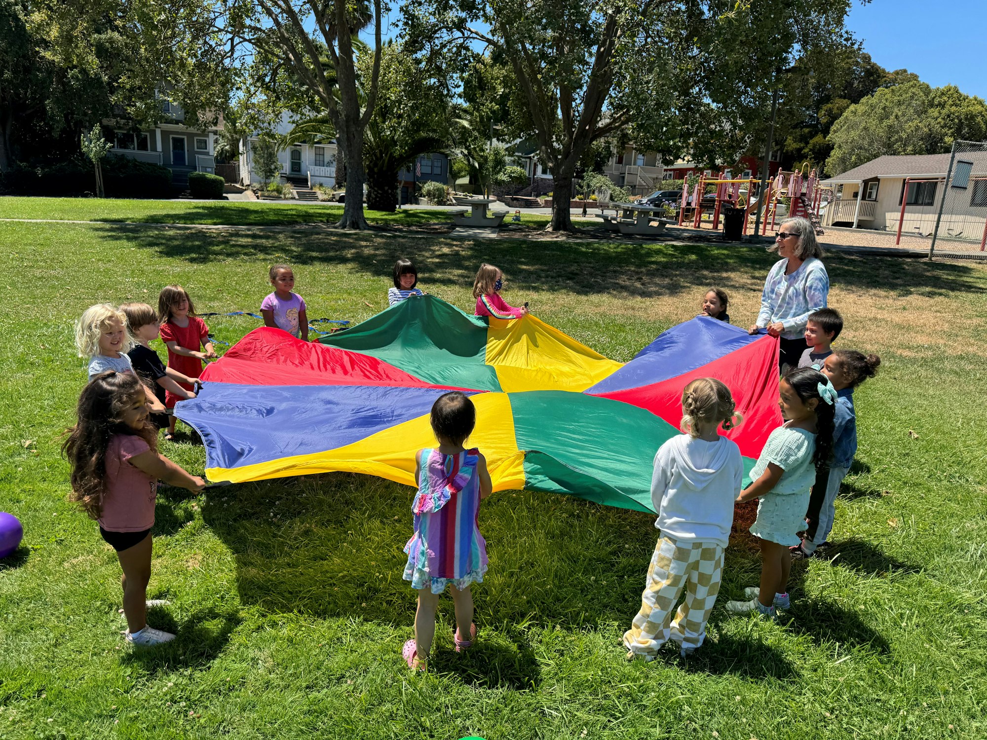 Children playing with a colorful parachute in a park with a playground in the background.