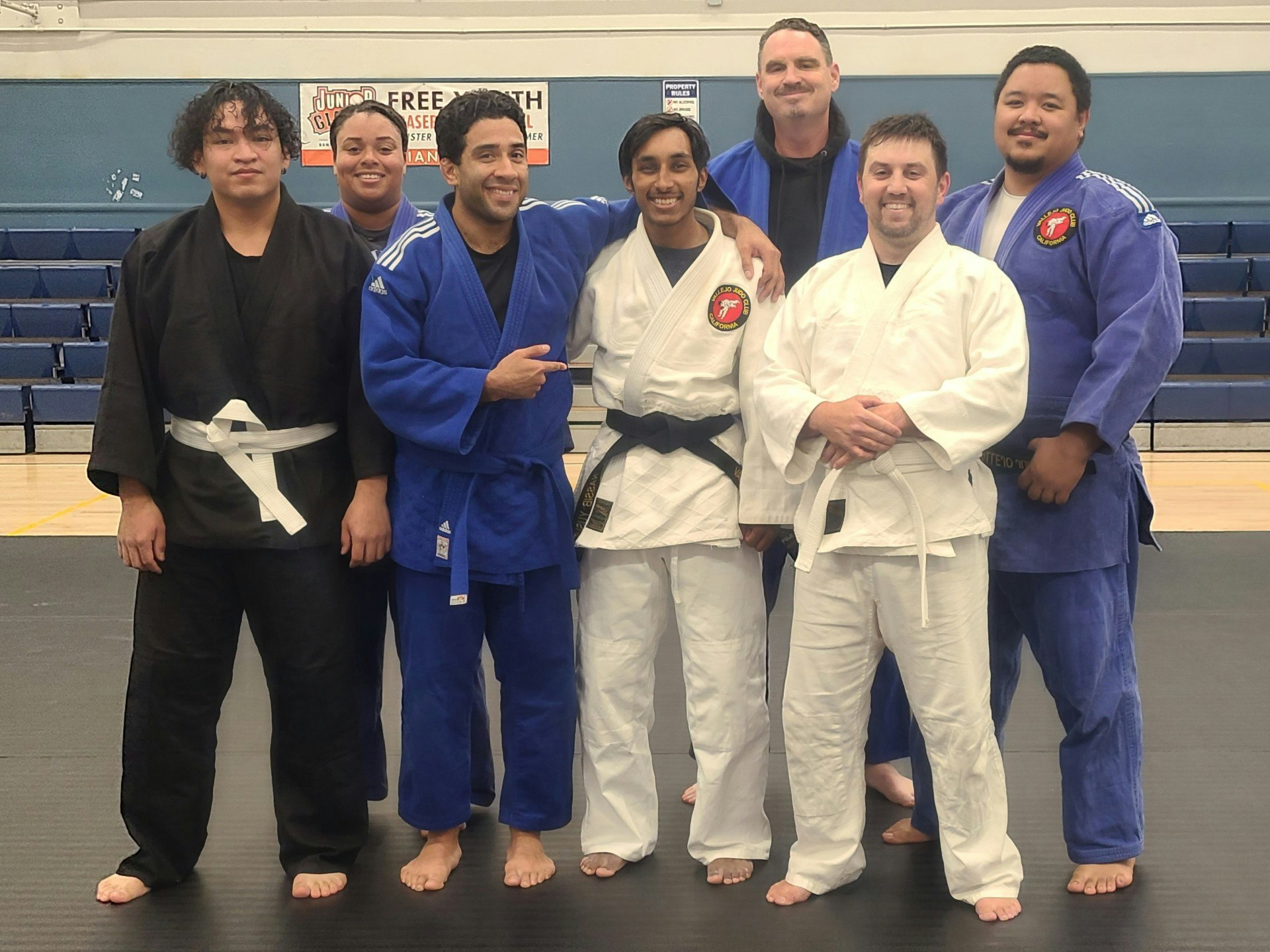 A group of seven people in martial arts uniforms, smiling on a training mat.