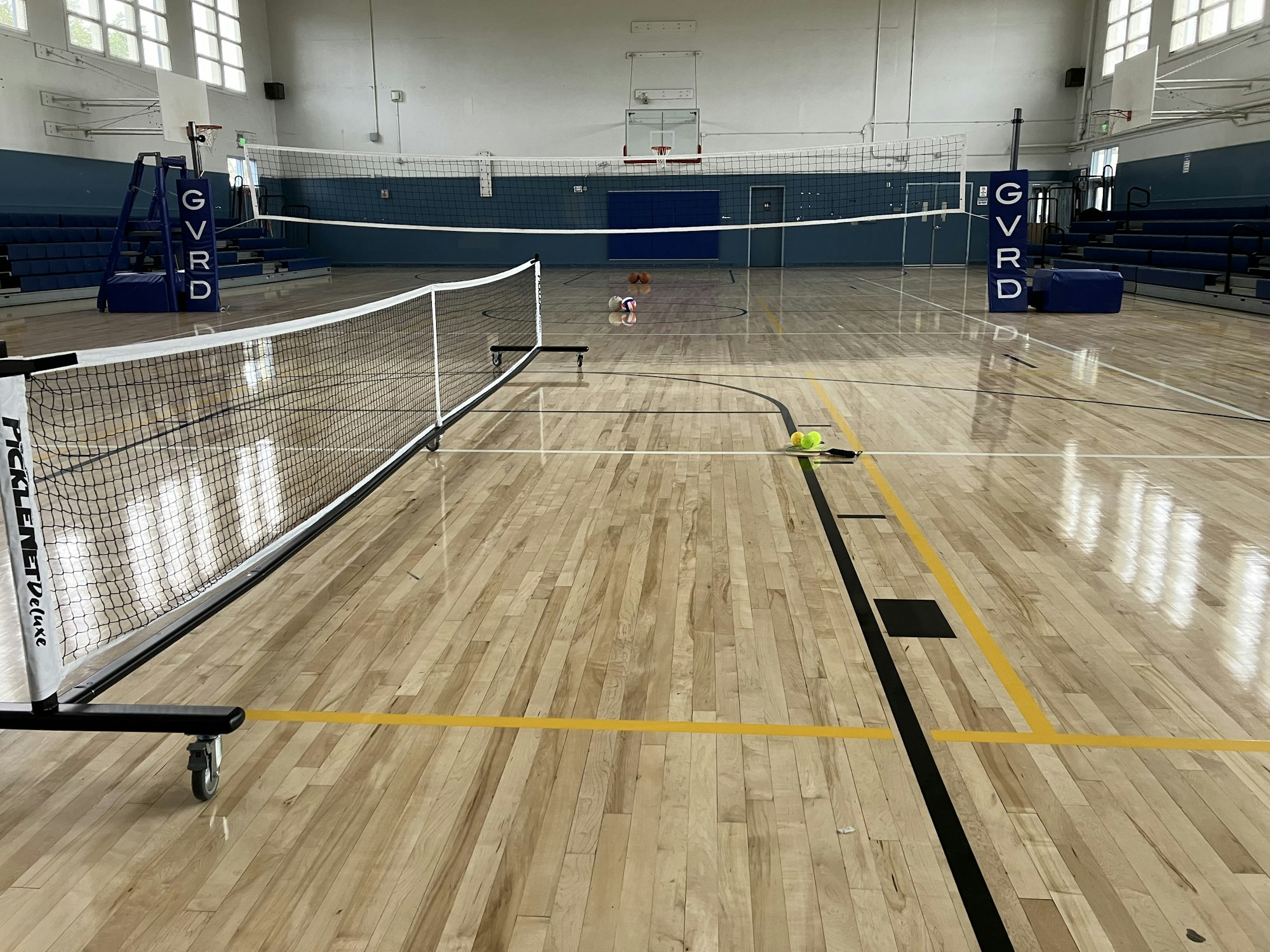 An indoor gym with a pickleball net set up, tennis balls on court, and volleyball equipment in the background.