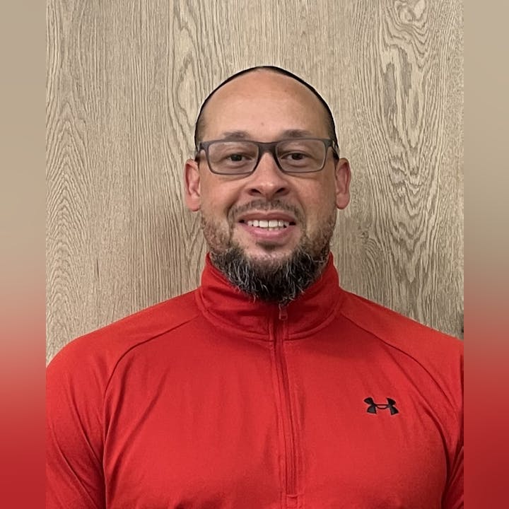 A person wearing glasses and a red Under Armour jacket stands against a wooden backdrop, smiling.