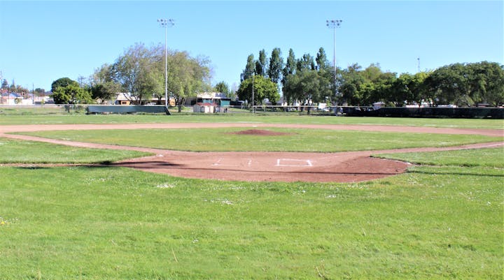 A baseball field with a dirt infield, grass outfield, and surrounding trees and structures under a clear blue sky.