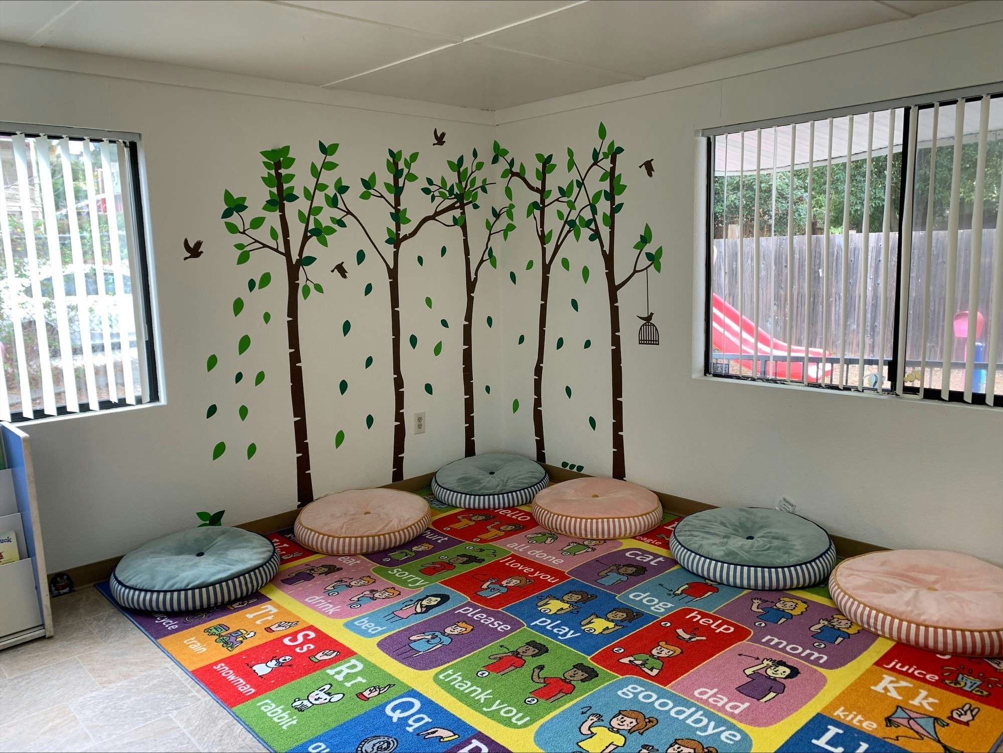 A children's playroom with wall decals of trees, birds, and cushions on the floor, and an educational alphabet rug.