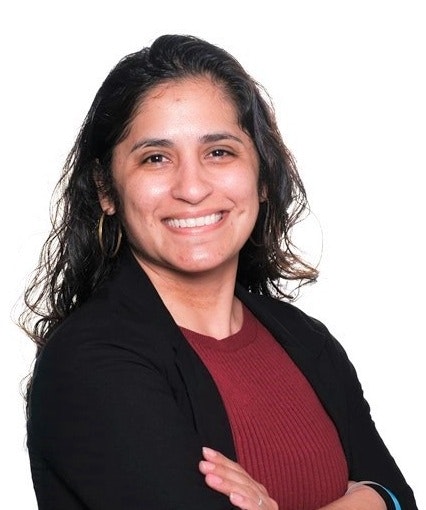 The image shows a smiling woman with long, curly hair, wearing a black blazer over a maroon top, standing with her arms crossed.