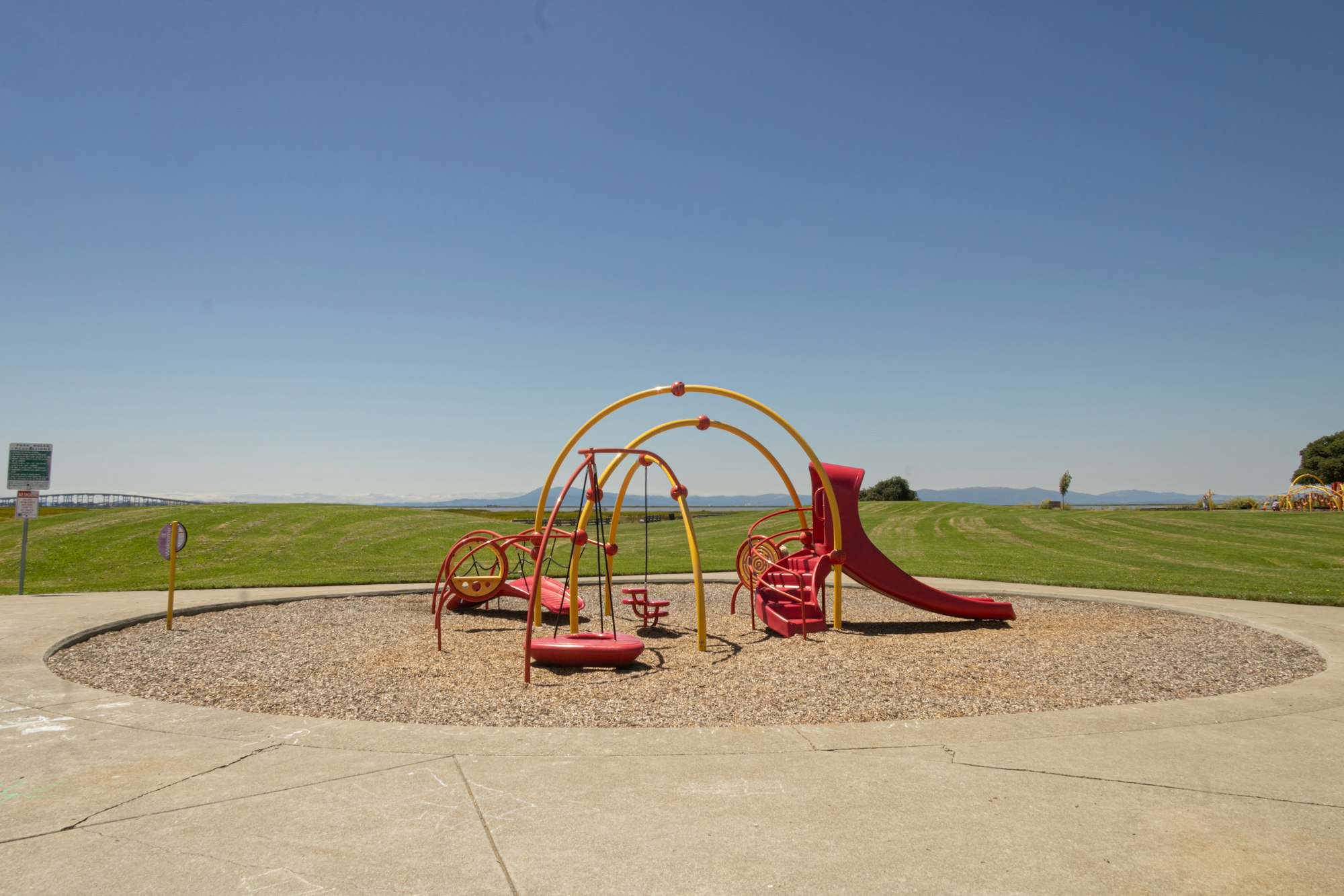 Red and yellow playground equipment with slides and swings on a sunny day, clear blue sky, grassy field in the background.