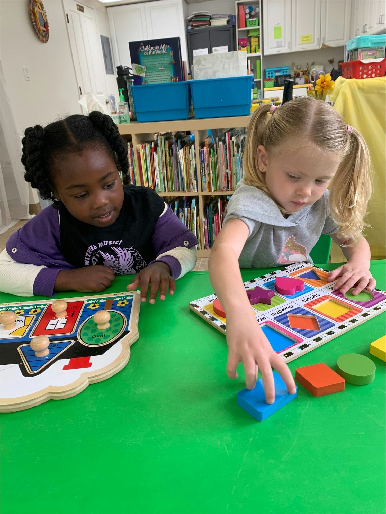 Two children engaged in a colorful shape-matching puzzle game, with educational books and materials in the background.