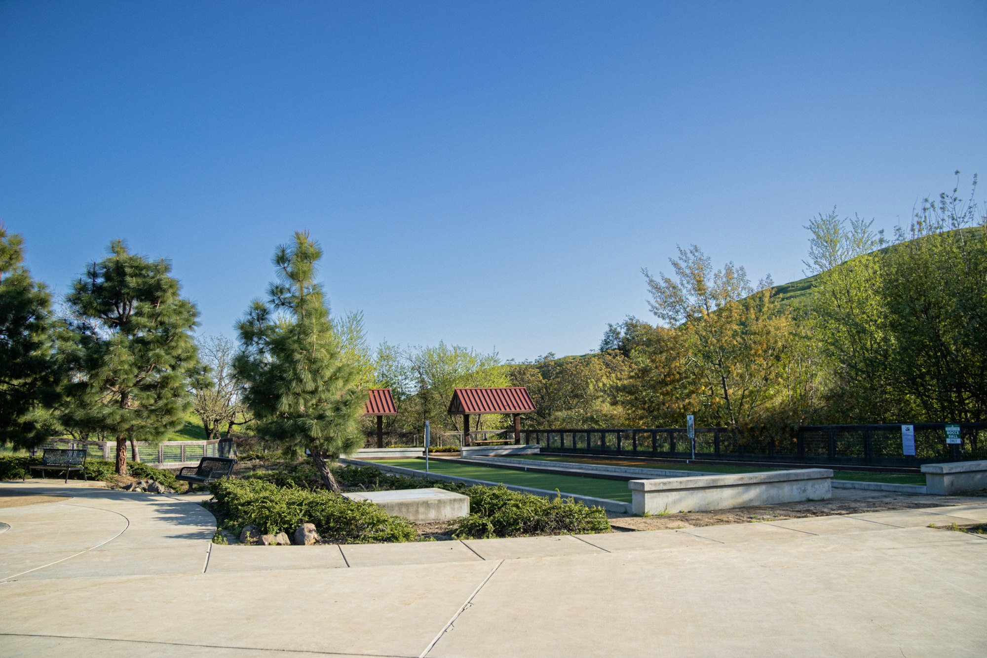 A tranquil park scene with bocce courts, benches, trees, and a blue sky.