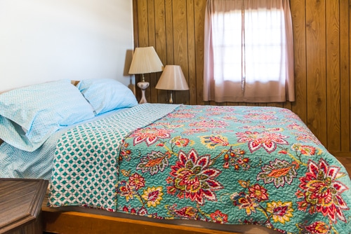 A cozy bedroom with a bed featuring a colorful quilt, light blue pillows, a wooden wall, a lamp, and a window with sheer curtains.