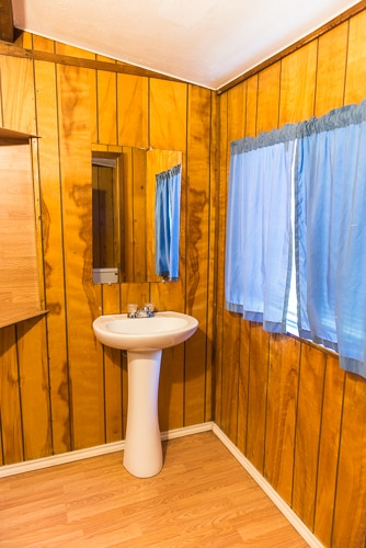 A small, wooden-paneled bathroom featuring a white sink, a mirror, and blue curtains over a window.