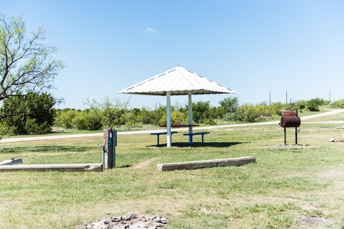 A picnic area features a covered table, grill, and grassy space under a clear blue sky. Ideal for outdoor gatherings.