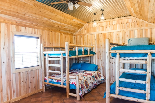 A cozy wooden room featuring two sets of bunk beds and a double bed, all with colorful bedding and natural lighting.