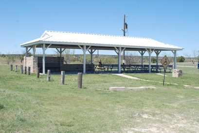 A covered picnic area with benches and tables, surrounded by grass and a few posts, under a clear blue sky.