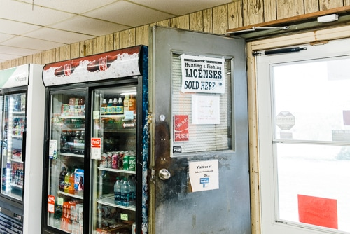 The image shows a convenience store entrance with signs about hunting and fishing licenses, and soda coolers nearby.