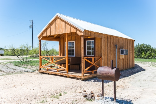 A small wooden cabin with a porch and a mailbox, set in a clear outdoor area under a blue sky.