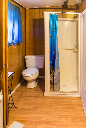 The image shows a small bathroom with a shower, toilet, wooden paneling, and a colorful shower curtain.