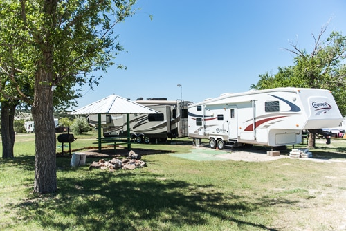 The image shows two RVs parked near a shaded gazebo and trees in a grassy outdoor area, suggesting a camping or picnic setting.