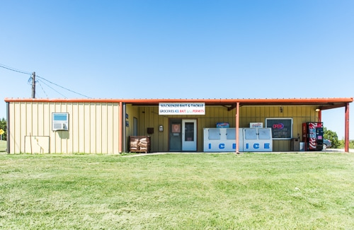 A simple country grocery store with a large sign, ice machine, and a vending machine in a grassy area under a clear blue sky.