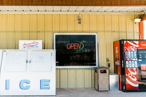 A storefront with an "OPEN" sign, a large ice cooler, a trash can, and a Coca-Cola vending machine.