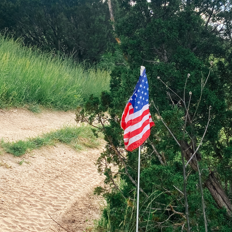 The image features a small American flag beside a dirt path surrounded by greenery.