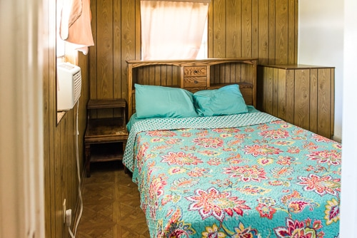 A cozy bedroom featuring a wooden bed frame, teal pillows, floral blanket, and window with light curtains.