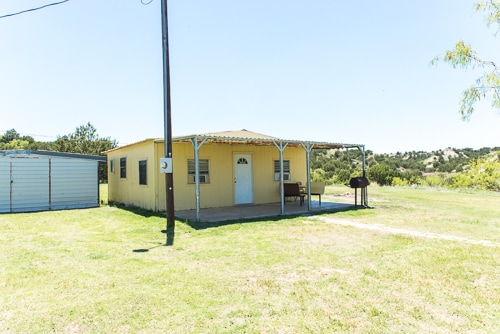 A yellow house with a porch sits on a grassy lot, with a nearby shed and a clear blue sky above.