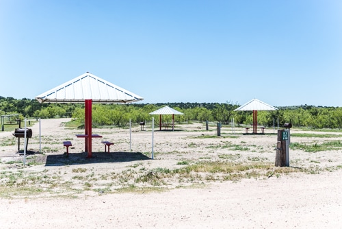 The image shows a picnic area with shaded tables, surrounded by sparse vegetation and open space under a clear blue sky.